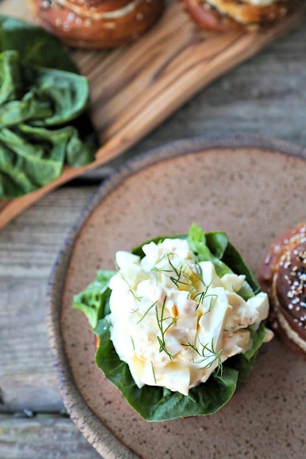 Dill Pickle Egg Salad on lettuce and pretzel roll, brown speckled pottery plate, lettuce leaves, wooden cutting board, rustic wood table, vertical