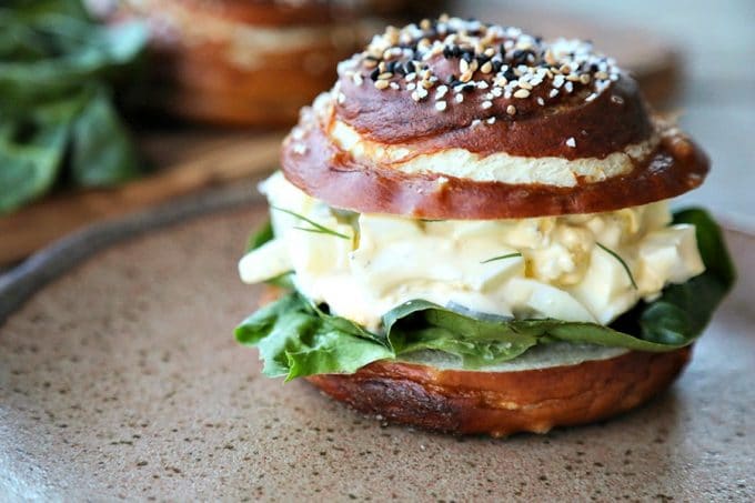 Dill Pickle Egg Salad on lettuce and pretzel roll, brown speckled pottery plate, lettuce leaves, wooden cutting board, rustic wood table, close up