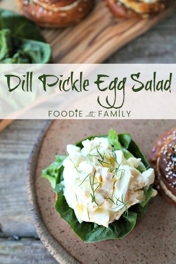 Dill Pickle Egg Salad with fresh dill on lettuce and soft pretzel roll, speckled brown pottery plate with rough edge, wooden cutting board with rolls and lettuce, rustic wooden table.