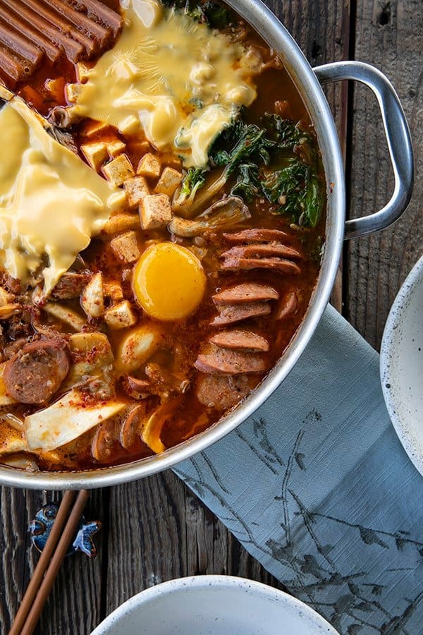 Korean Army Stew or Budae Jjigae with egg yolks and American cheese in a stainless steel everyday pan. Blue linen on the table with two white bowls and wooden chopsticks.