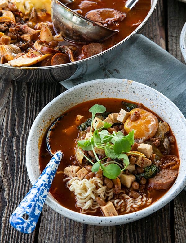 Korean Army Stew or Budae Jjigae in a white bowl with an egg yolk and a blue enamel spoon.