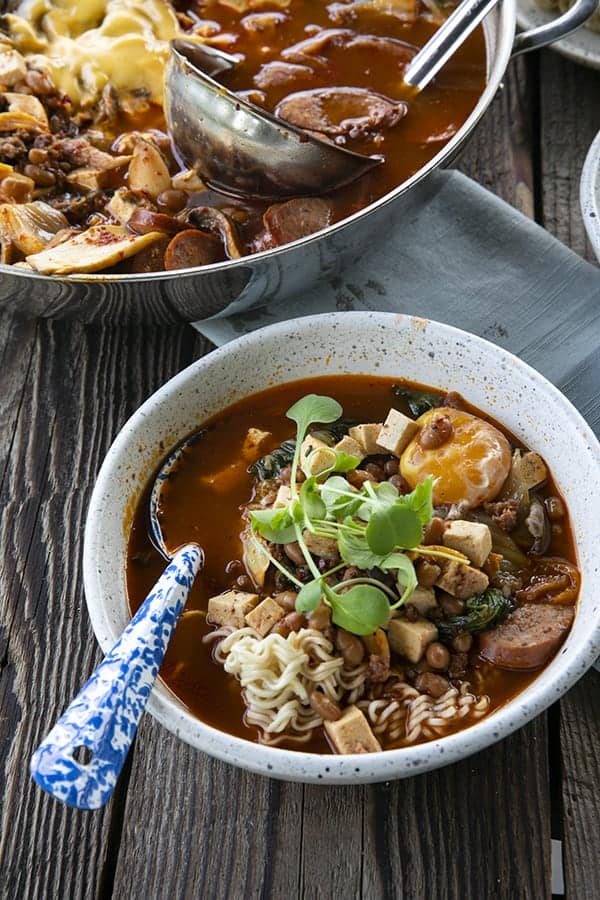 Korean Army Stew or Budae Jjigae in a white bowl with an egg yolk and a blue enamel spoon.