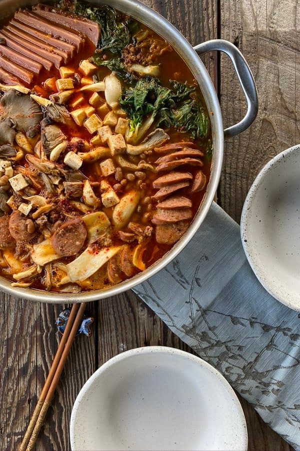 Korean Army Stew in a stainelss steel pan with two empty bowls, chopsticks, and a blue linen.