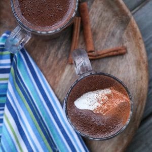 Blender made Spiced Hot Chocolate topped with cream and cinnamon, wooden tray, blue green and white towel, cinnamon sticks,